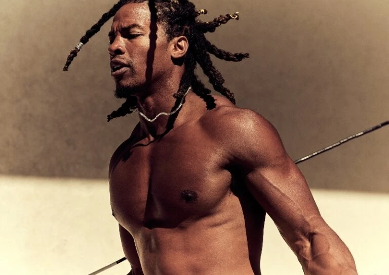 Toned and muscular Black man with dreadlocks gazes off-camera, wearing a delicate necklace against a neutral background, holding a spear.