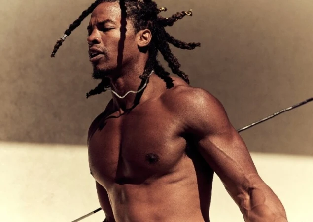 Toned and muscular Black man with dreadlocks gazes off-camera, wearing a delicate necklace against a neutral background, holding a spear.
