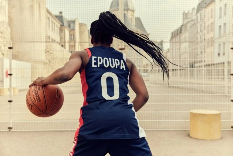 Woman in basketball uniform dribbling on an outdoor court in front of a tan building with round yellow cylinder prop beside her.