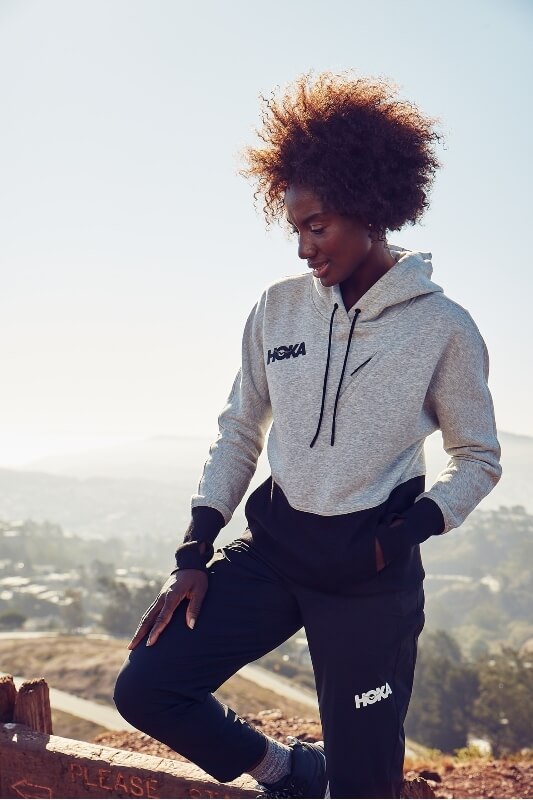 Athletic African-American woman in Hoka-branded activewear stands outside against a blurred cityscape background on a sunny day.