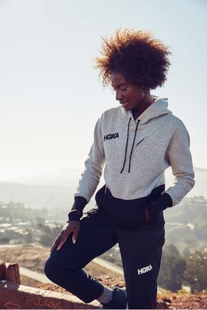Athletic African-American woman in Hoka-branded activewear stands outside against a blurred cityscape background on a sunny day.