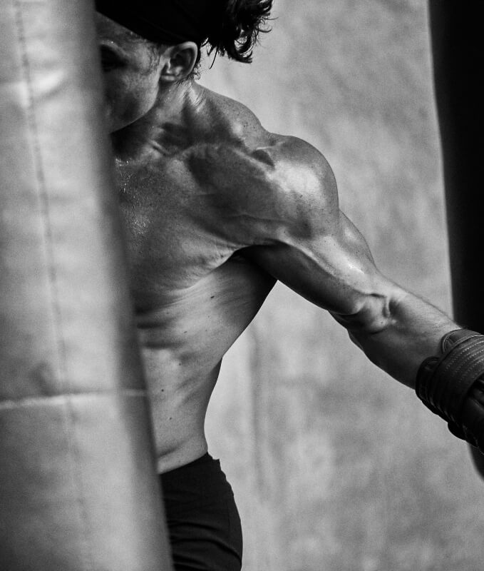 Monochrome shot of a muscular adult male wearing boxing gloves and a head band punching a punching bag in a gym setting.