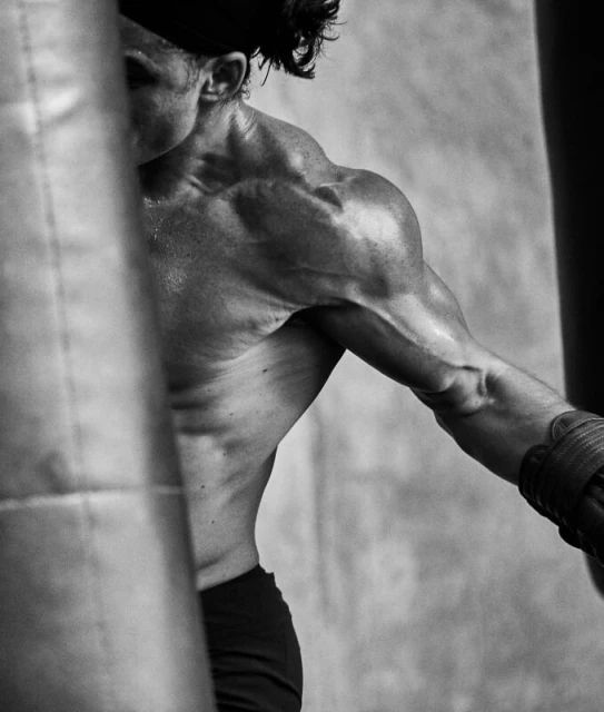 Monochrome shot of a muscular adult male wearing boxing gloves and a head band punching a punching bag in a gym setting.