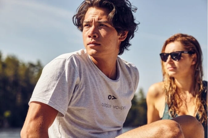 A young man in a white graphic t-shirt sitting outdoors with a woman wearing sunglasses in the background.