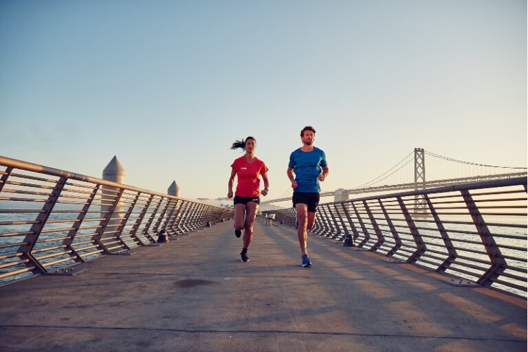 Two athletes, an adult woman and adult man, run on a concrete pier with metal railings, with a view of a bridge in the background.