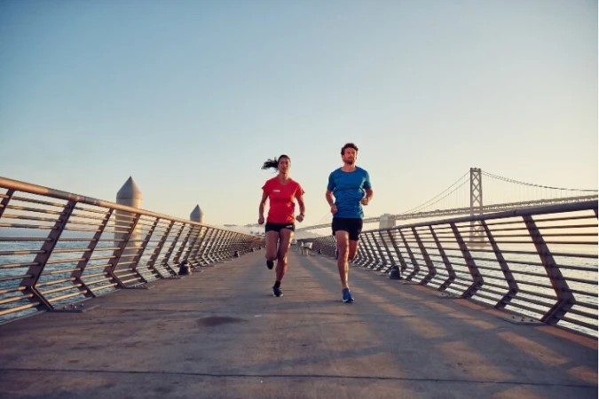 Two athletes, an adult woman and adult man, run on a concrete pier with metal railings, with a view of a bridge in the background.