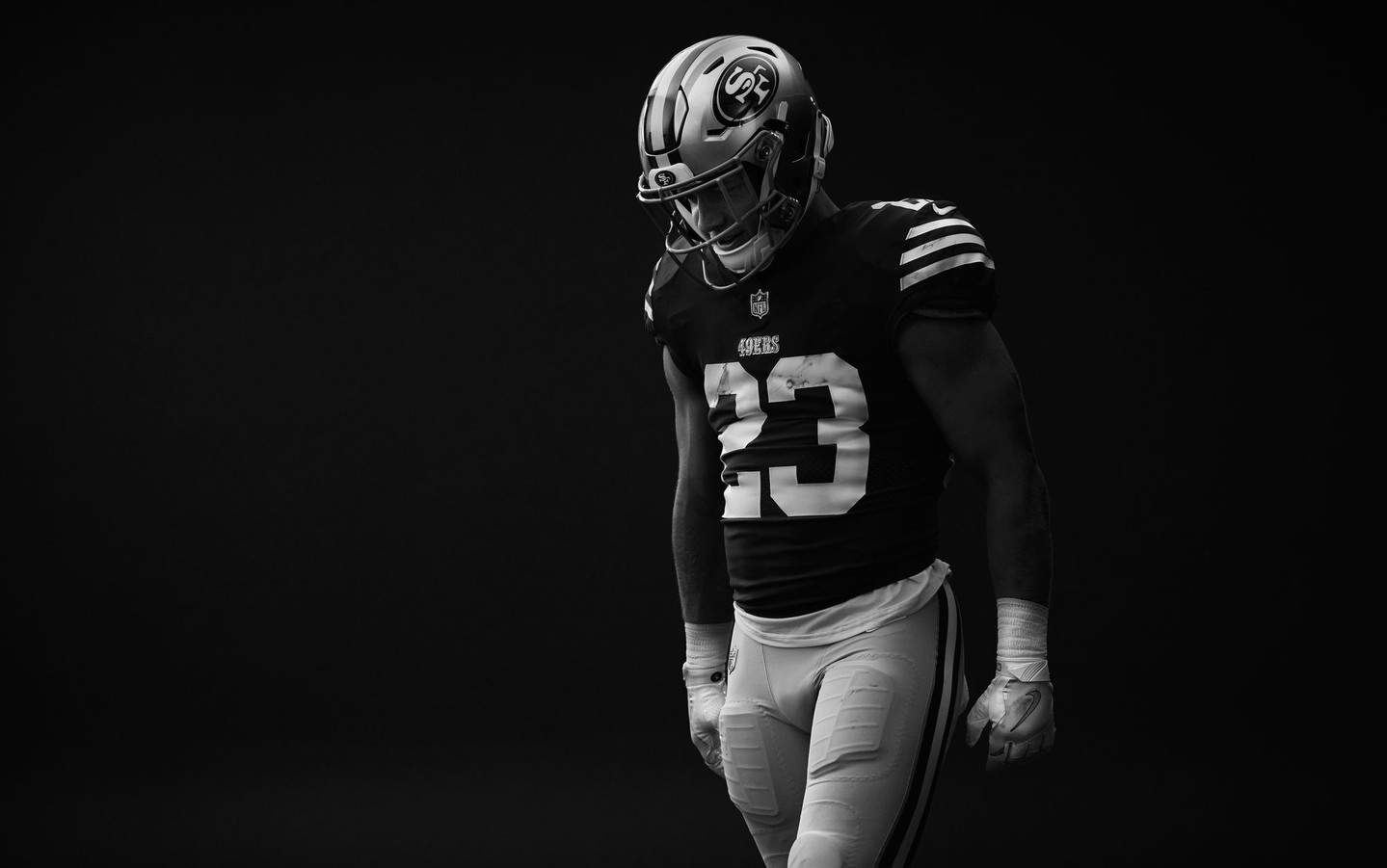 Monochrome portrait of an adult male football player in uniform with helmet, pads, and gloves standing against a dark background.
