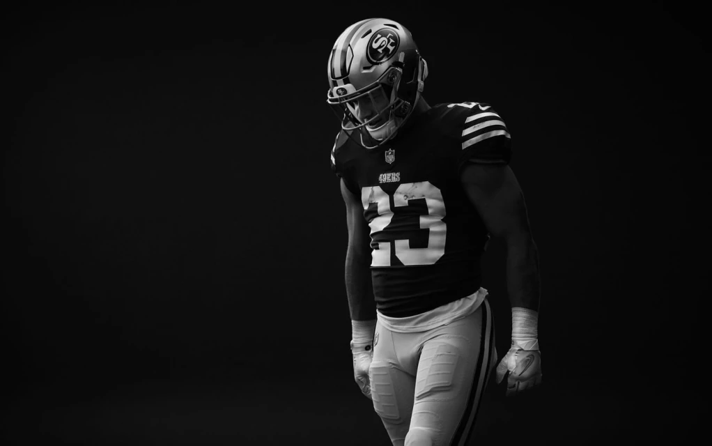 Monochrome portrait of an adult male football player in uniform with helmet, pads, and gloves standing against a dark background.