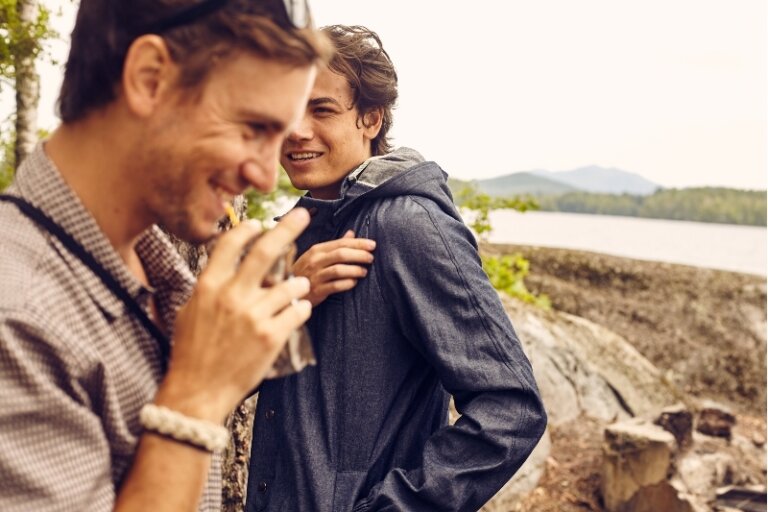 Two men are standing near a lake surrounded by mountains and trees. One is drinking from a carton. They are dressed casually.