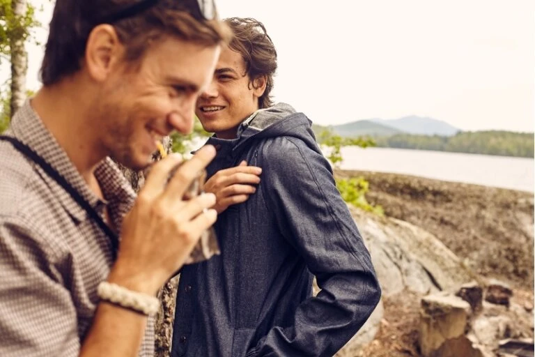 Two men are standing near a lake surrounded by mountains and trees. One is drinking from a carton. They are dressed casually.