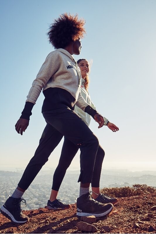 Two women walking uphill on a dirt path with city and ocean visible in the background distance under a bright sky