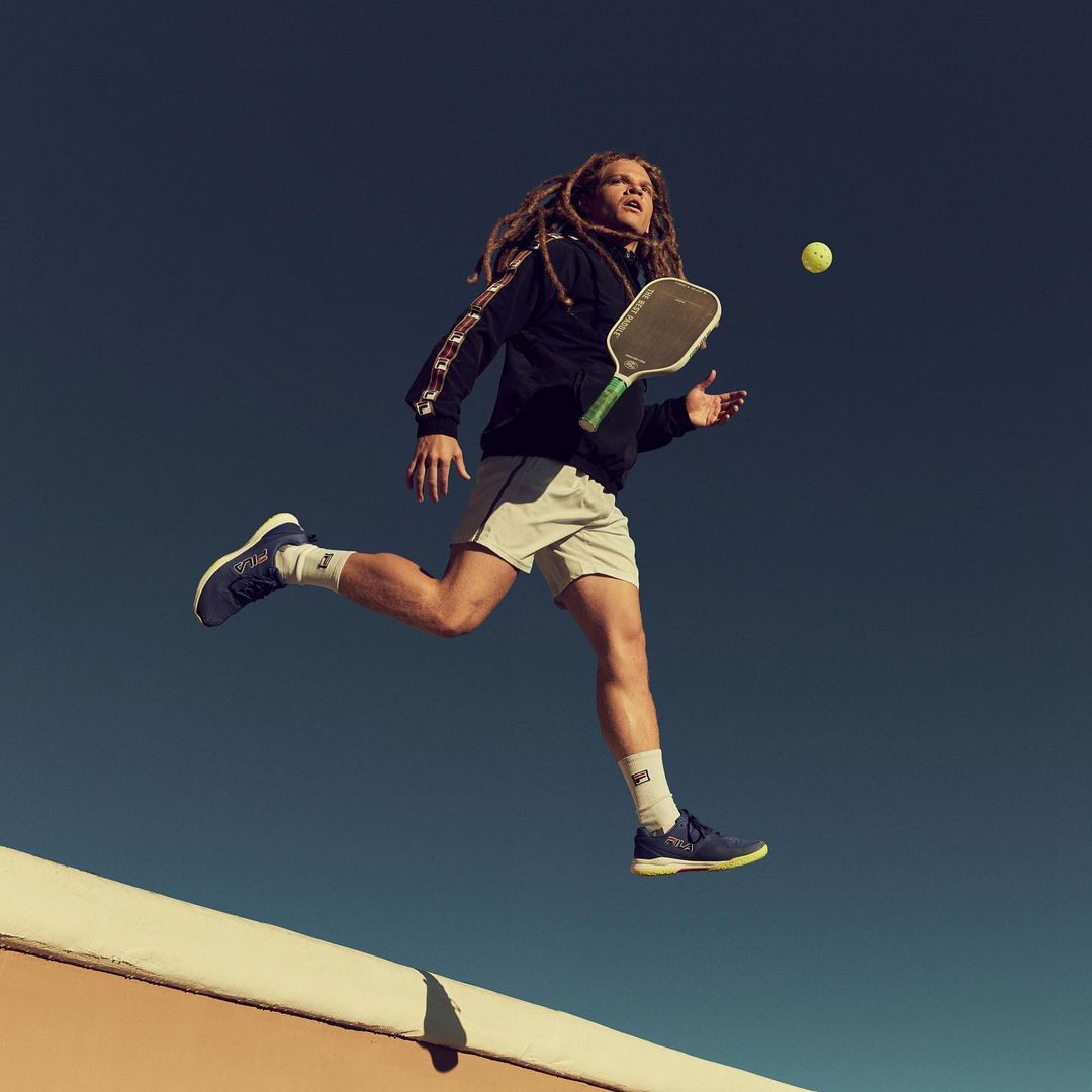 A male athlete with dreadlocks jumps in mid-air, holding a pickleball paddle and ready to hit a yellow ball against a dark blue sky.