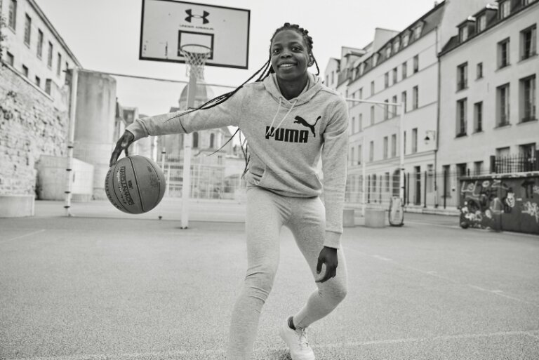 Young Black woman in Puma sweats dribbling a basketball on an outdoor court, urban setting, black and white.