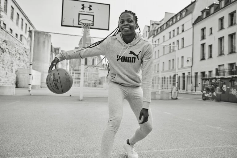 Young Black woman in Puma sweats dribbling a basketball on an outdoor court, urban setting, black and white.