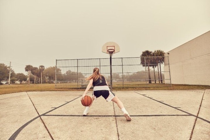 Female basketball player dribbling on an outdoor court with a hoop and a cloudy sky in the background, capturing a moment of athletic practice.