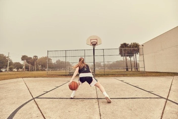 Female basketball player dribbling on an outdoor court with a hoop and a cloudy sky in the background, capturing a moment of athletic practice.
