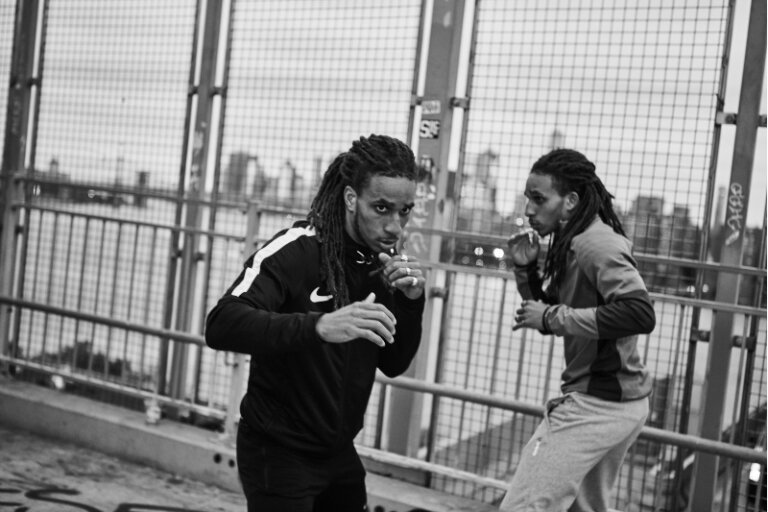 Two athletic men practice boxing on a city bridge with a distant skyline, showcasing fitness and urban training in black and white.