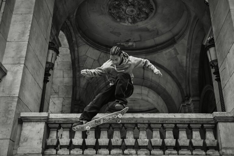 Monochrome image of a skateboarder performing a trick in front of an arched stone building with decorative railings and vintage lamps.