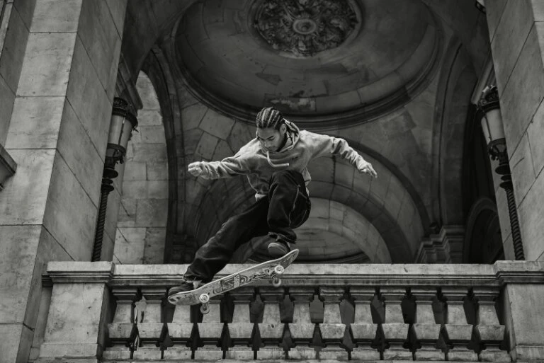 Monochrome image of a skateboarder performing a trick in front of an arched stone building with decorative railings and vintage lamps.