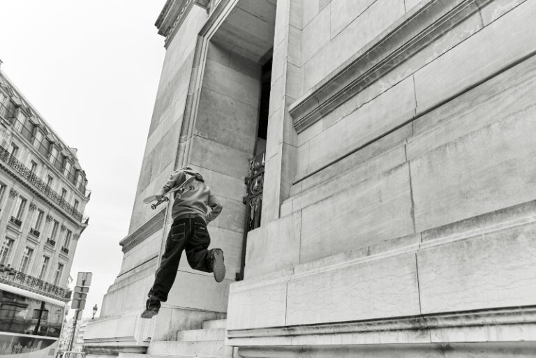 Black and white image of a child jumping off steps holding a skateboard, with urban architecture and sky in the background.