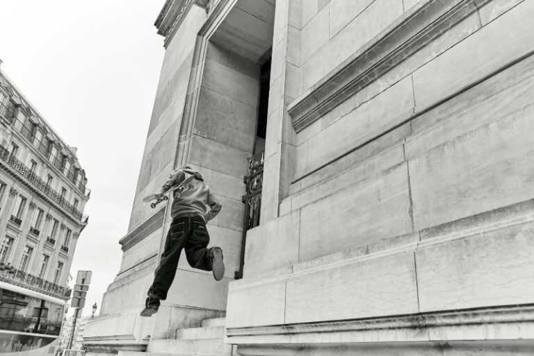 Black and white image of a child jumping off steps holding a skateboard, with urban architecture and sky in the background.