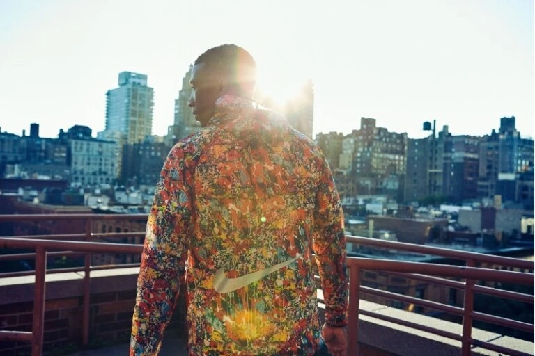 Adult man wearing colorful jacket stands on rooftop with city skyline in the background during daytime under bright sunlight.