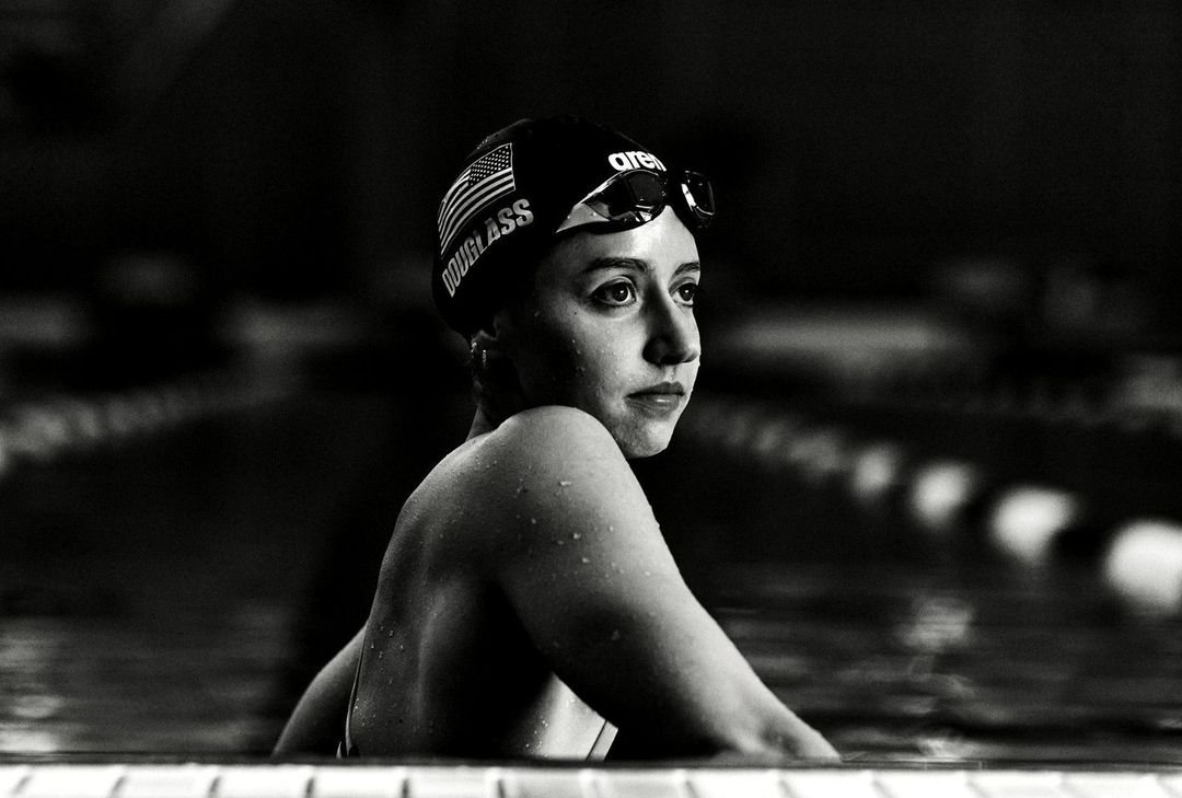 Black and white shot of a young adult female swimmer at the edge of a pool wearing a swim cap and goggles with a determined expression.