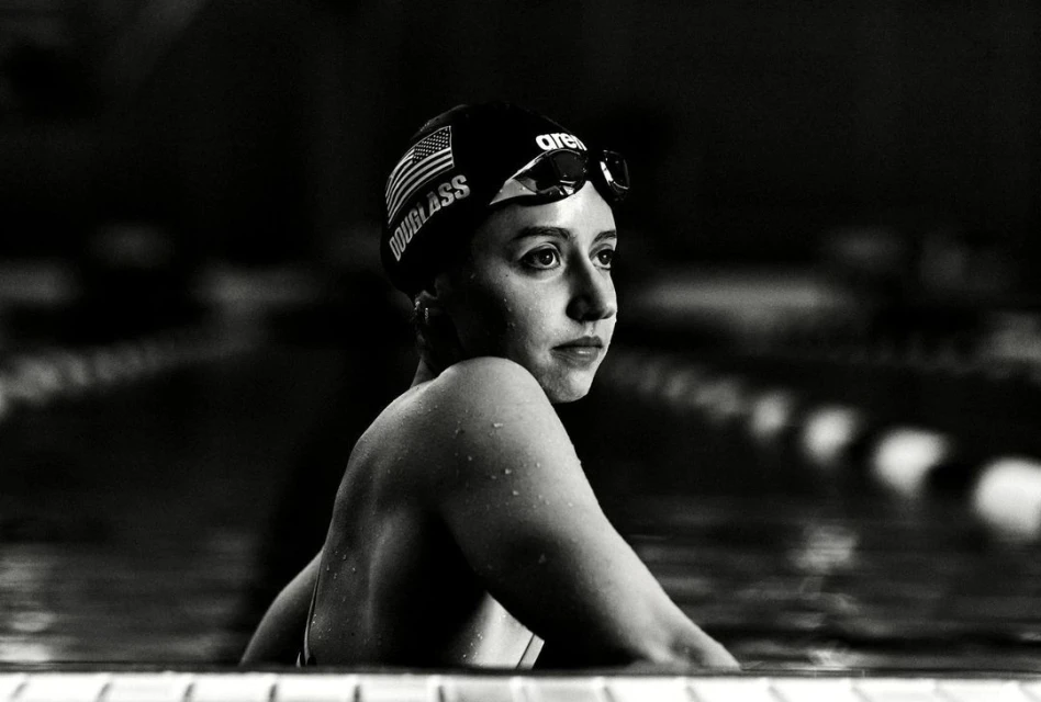 Black and white shot of a young adult female swimmer at the edge of a pool wearing a swim cap and goggles with a determined expression.