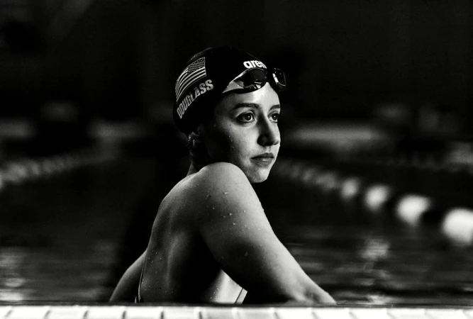 Black and white shot of a young adult female swimmer at the edge of a pool wearing a swim cap and goggles with a determined expression.