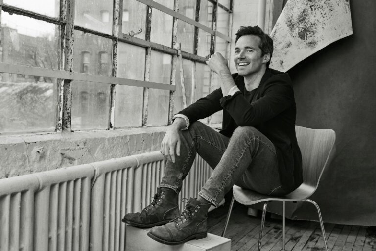 Monochrome portrait of a man sitting by a window in a jacket and jeans with combat boots, looking off into the distance with a smile.