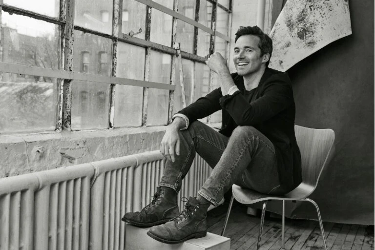 Monochrome portrait of a man sitting by a window in a jacket and jeans with combat boots, looking off into the distance with a smile.