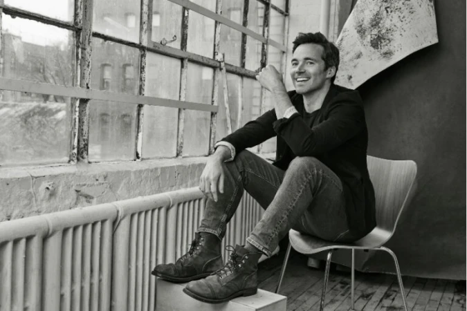 Monochrome portrait of a man sitting by a window in a jacket and jeans with combat boots, looking off into the distance with a smile.