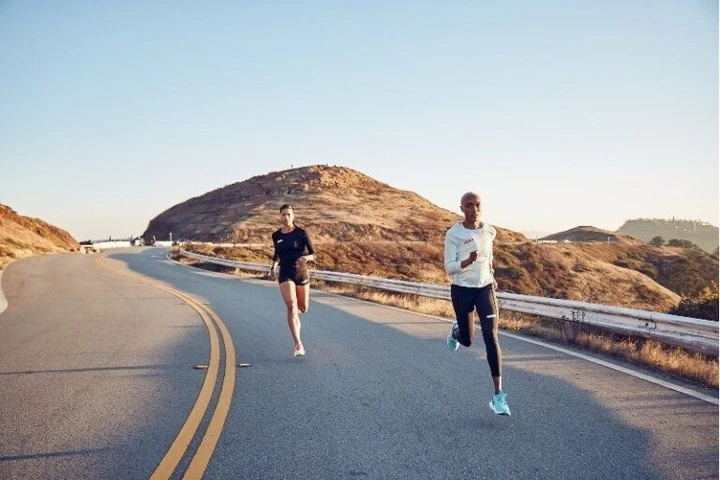 Two athletic women run on a curving asphalt road, with a grassy hill and blue sky in the background under bright sunlight, shot at eye-level.