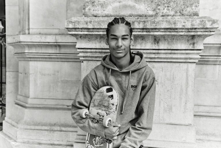 Young man with braided hair in gray hoodie holding a skateboard, poses outdoors in front of a stone pillar, black and white image.