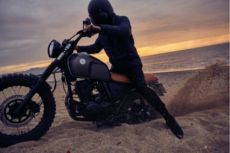 Motorcycle rider on a beach at dusk. The rider wears a helmet and dark clothing, with soft sunset colors in the sky.