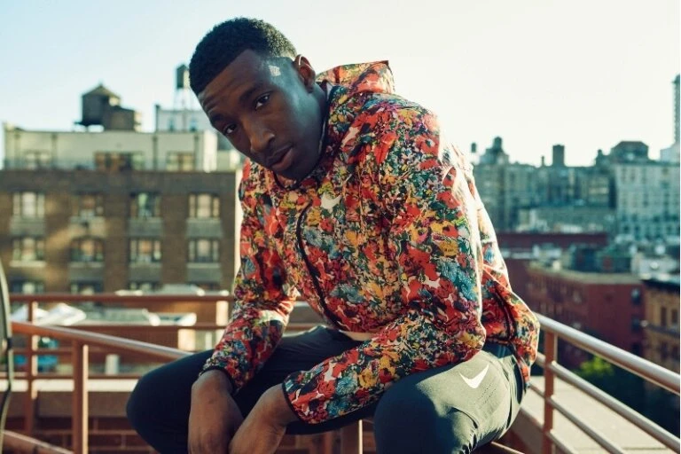 Young man in floral hoodie crouches on rooftop overlooking city buildings on a sunny day, wearing athletic pants with the Nike logo.