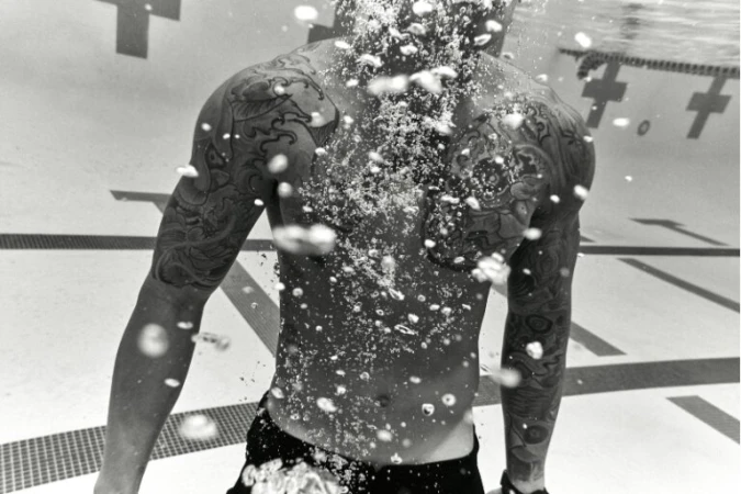 Black and white underwater portrait of a tattooed man swimming in a pool by Coty Tarr.