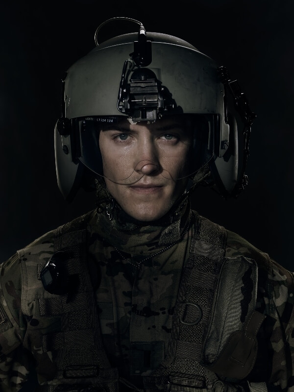 Close-up of a young adult male wearing a military helmet and camouflage uniform against a dark background in a studio shot.