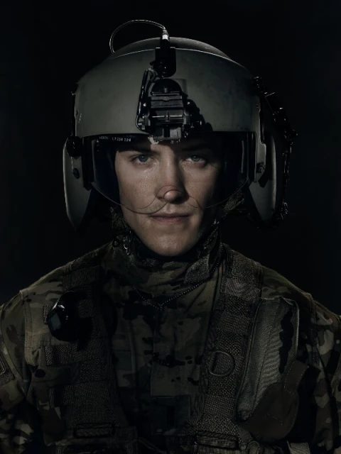 Close-up of a young adult male wearing a military helmet and camouflage uniform against a dark background in a studio shot.