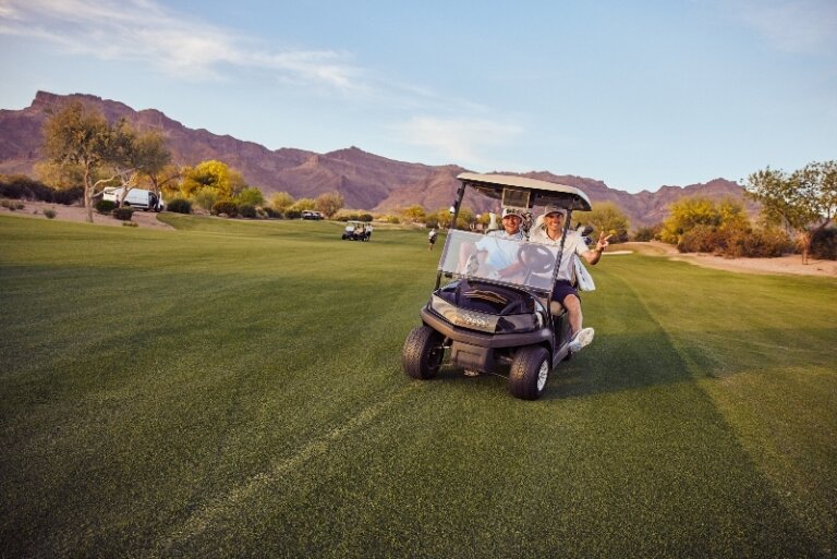 Two smiling adult golfers in a golf cart on a lush green course with mountains in the background under a clear sky during daytime.