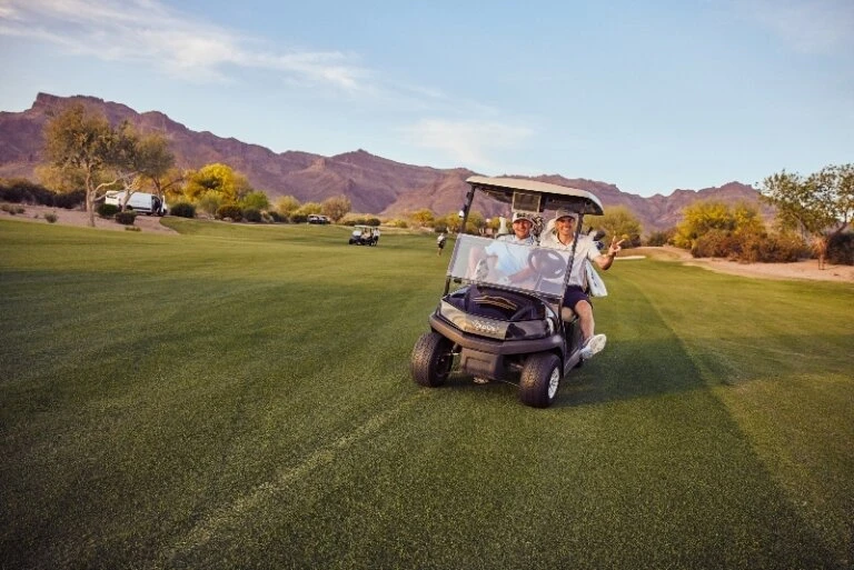 Two smiling adult golfers in a golf cart on a lush green course with mountains in the background under a clear sky during daytime.