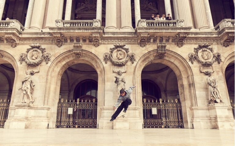 Adult male skateboarder in mid-air performing a trick in front of a grand stone building with arches and statues in daytime.