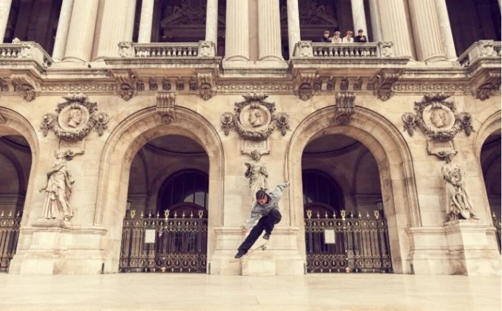 Adult male skateboarder in mid-air performing a trick in front of a grand stone building with arches and statues in daytime.