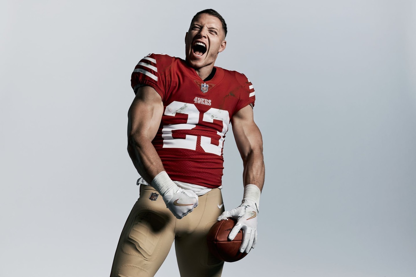 A muscular football player in a 49ers uniform, number 23, shouts with clenched fist and holding a football against a grey background.