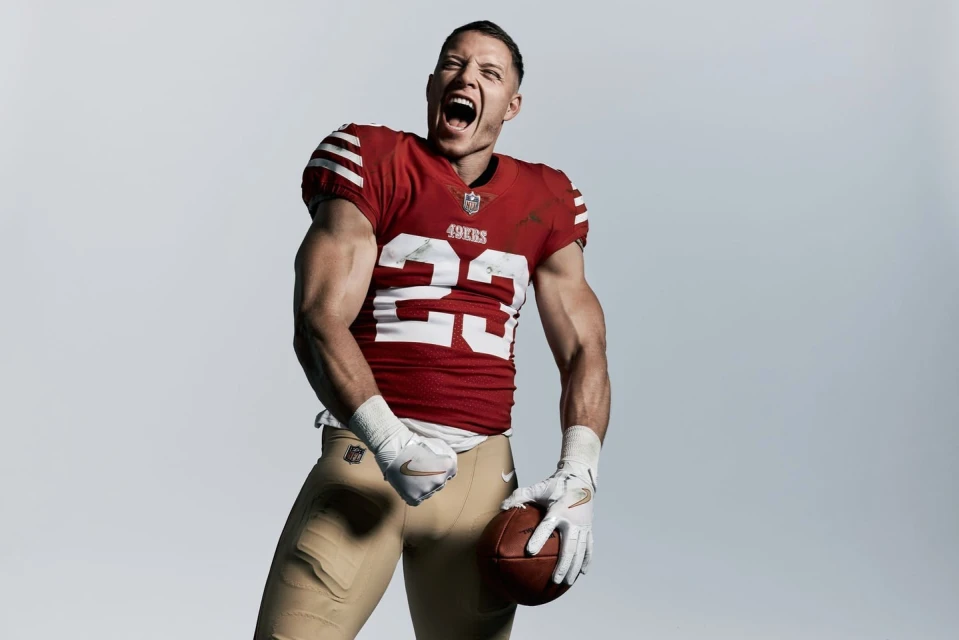 A muscular football player in a 49ers uniform, number 23, shouts with clenched fist and holding a football against a grey background.