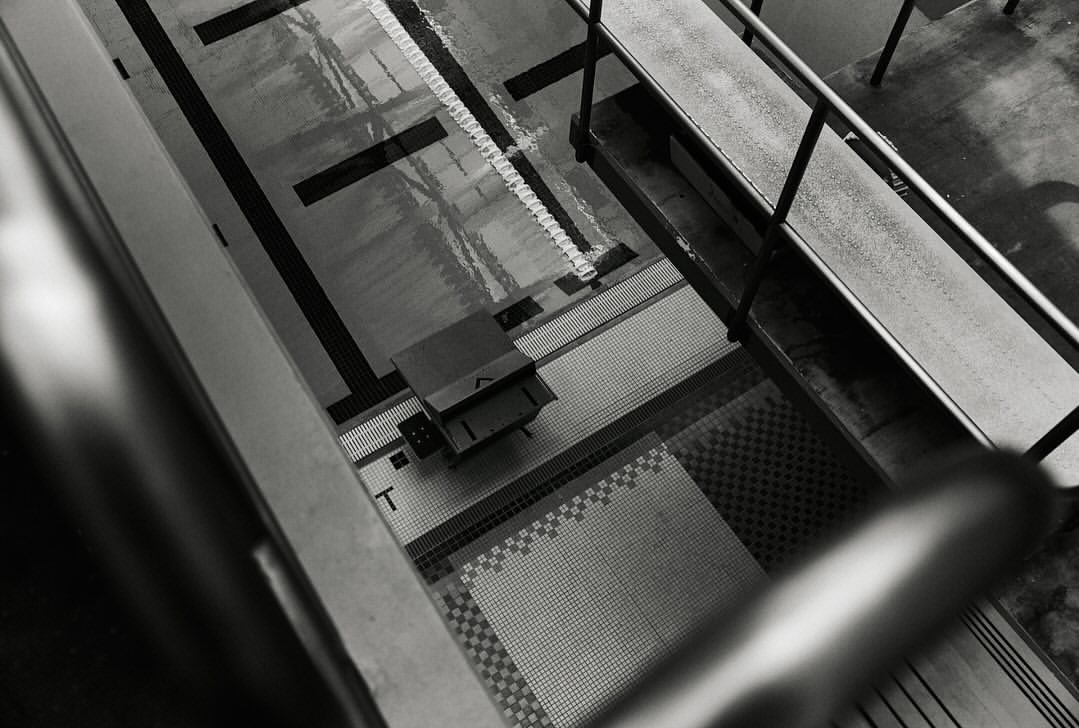 High-angle, black and white photo of an indoor swimming pool with a diving block, water, and tiled pool deck.