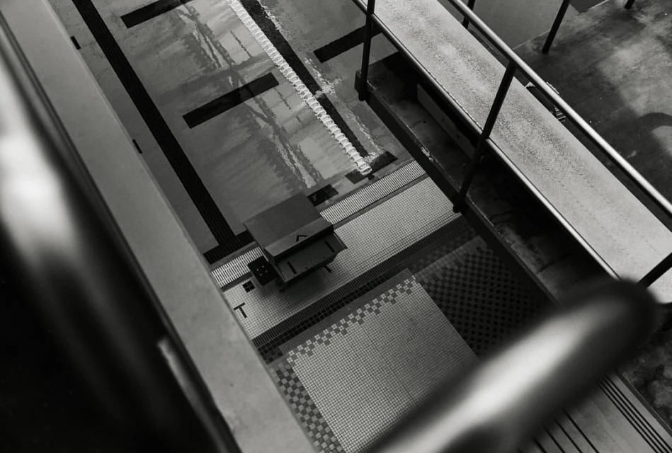 High-angle, black and white photo of an indoor swimming pool with a diving block, water, and tiled pool deck.