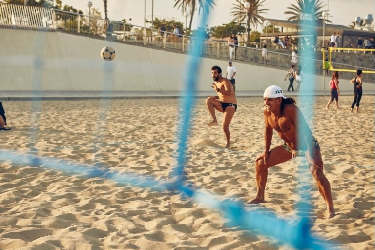 Two shirtless men play beach soccer on a sunny day with a blue net and several people in the background on sandy beach near an urban boardwalk.