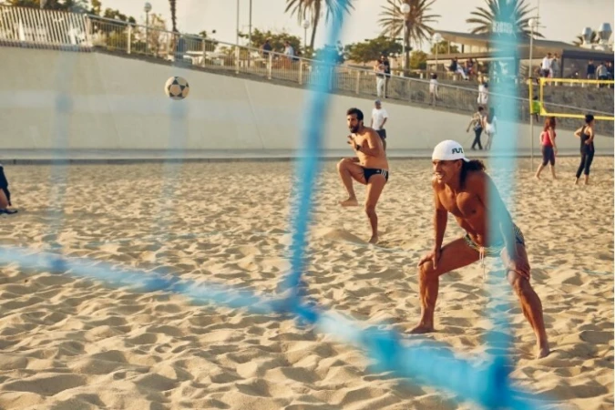 Two shirtless men play beach soccer on a sunny day with a blue net and several people in the background on sandy beach near an urban boardwalk.