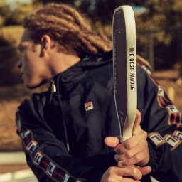 Close-up of a person holding a pickleball paddle, wearing a dark athletic jacket with striped sleeves, outdoors.